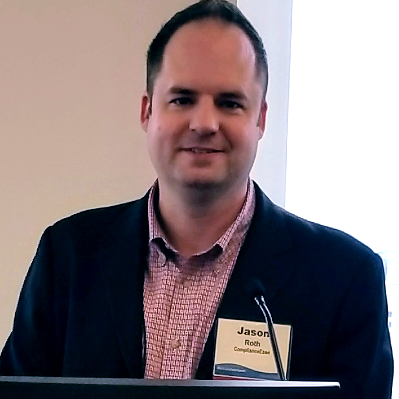 Jason Roth, wearing a dark suit jacket and checkered shirt and a name tag that reads 'Jason' is standing behind a podium
