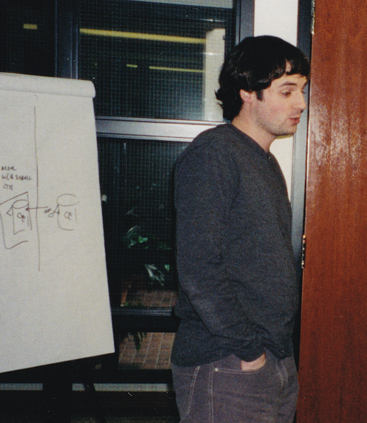 In his early career at his startup offices, Jason Roth, with dark curly hair and wearing a dark sweater, stands next to a large paper flipchart with diagrams drawn on it